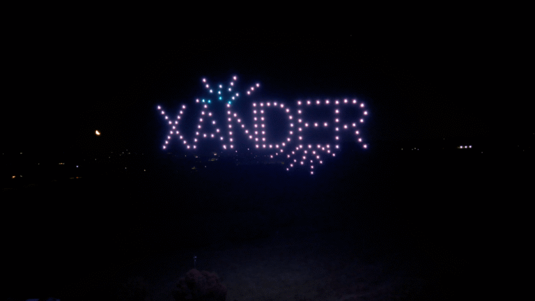 Drones spelling “XANDER” in pale pink and white lights, with turquoise and pink light bursts in the night sky.