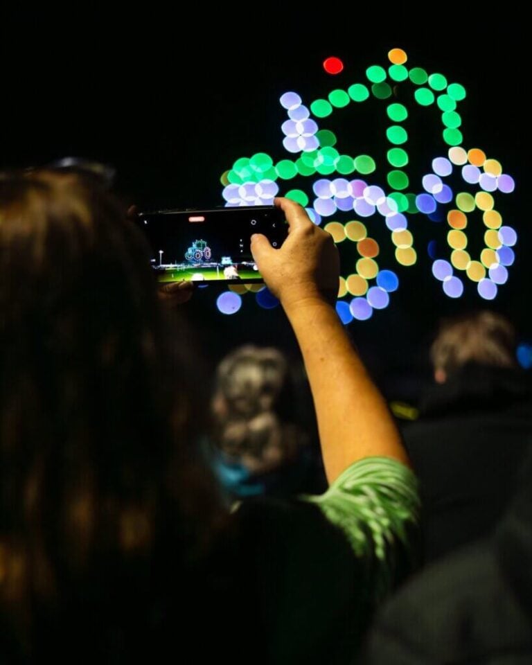 A spectator films a green tractor drone formation on a phone, with colourful lights filling the night sky behind it.